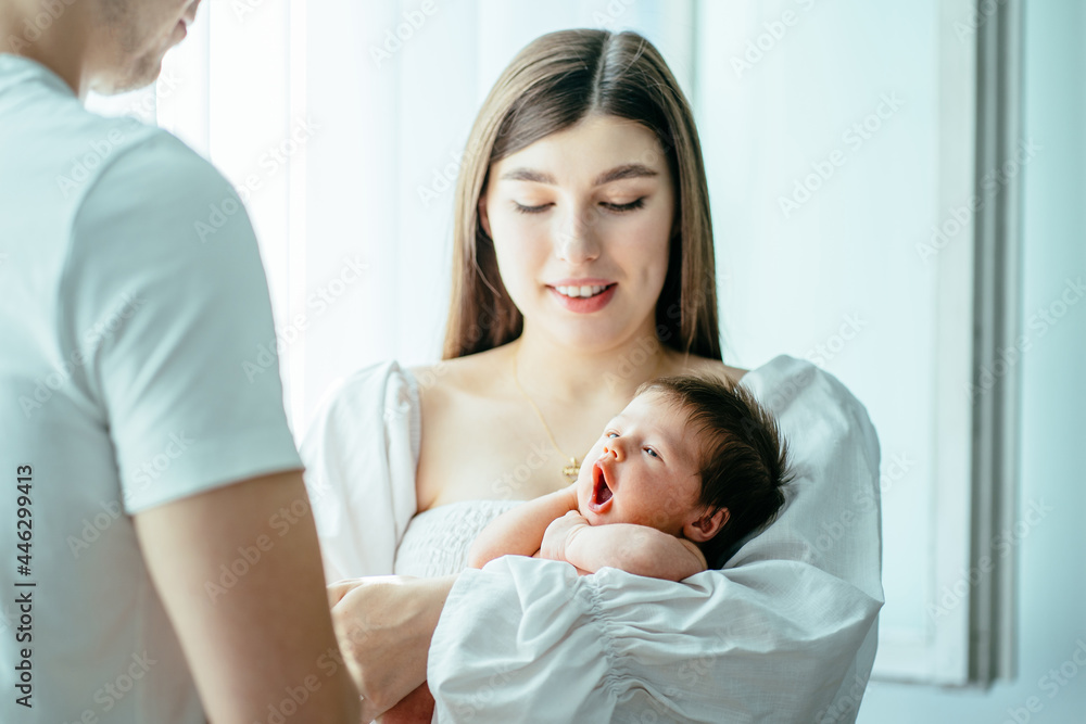 Obraz premium Attractive mother holding cute newborn baby boy in her arms near the window. Cute sleepy baby girl yawning. Happy dad and mom caring for little infant child together.