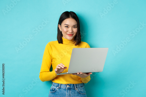 Young asian woman freelancer working on laptop and smiling, standing with computer over blue background photo