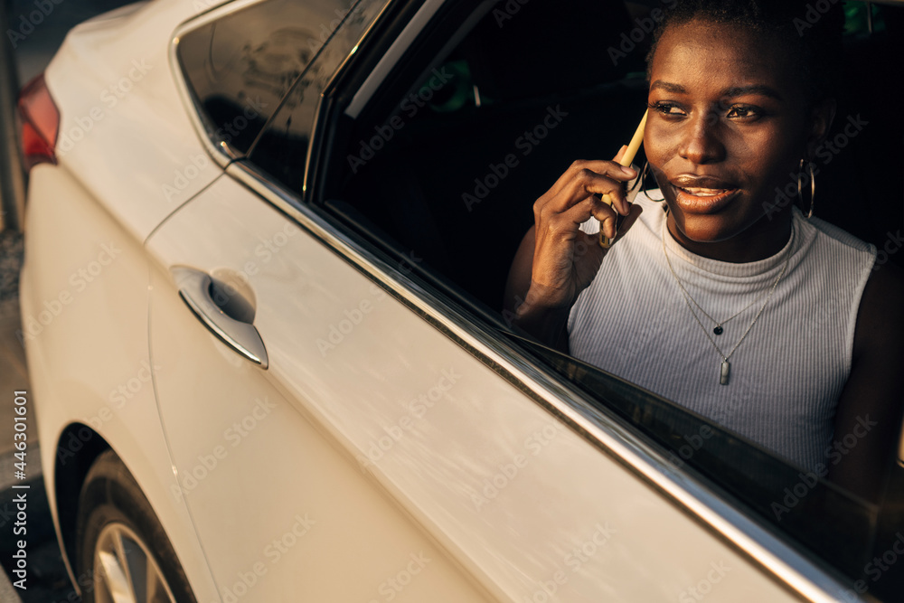 Naklejka premium African American girl talking on smartphone while sitting in the back seat of the car, looking out the window.