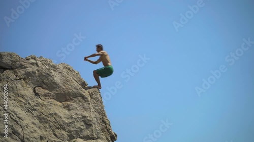 A young man jumping from a high cliff into the sea doing a back somersault on a hot summer day close-up.