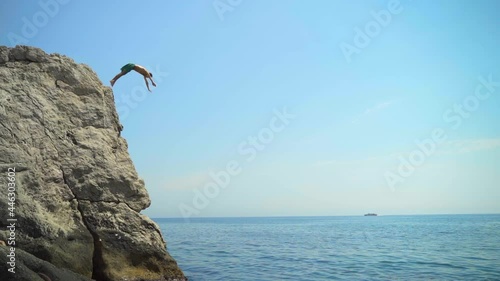 A young man jumping from a high cliff into the sea doing a forward somersault in slow motion on a hot summer day