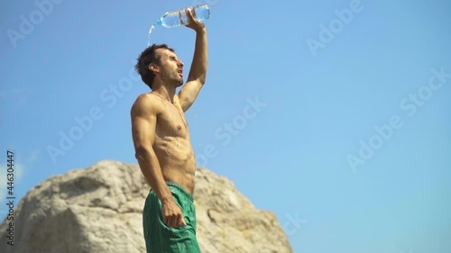 A young guy of athletic build in the heat opens a plastic bottle and pours fresh water on himself. The guy is doused with water from the heat in slow motion