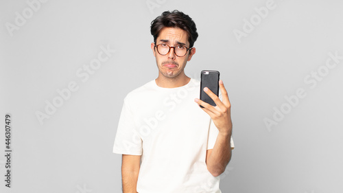 young hispanic man feeling sad and whiney with an unhappy look and crying and holding a smartphone photo