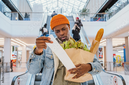 Surprised African-American man in denim jacket looks at receipt total in sales check holding paper bag with products in mall photo