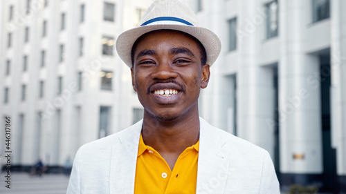 Black successful businessman in yellow t shirt and white suit jacket walks along grey street and smiles photo