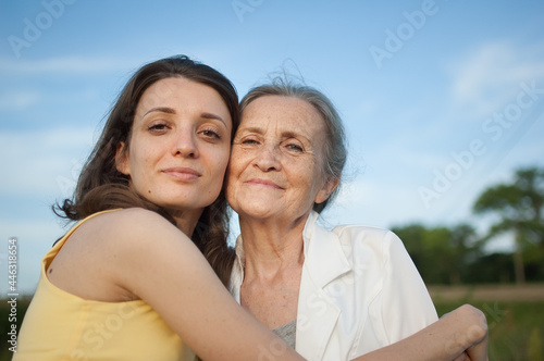Senior mother with gray hair with her adult daughter looking at the camera in the garden and hugging each other during sunny day outdoors, mothers day