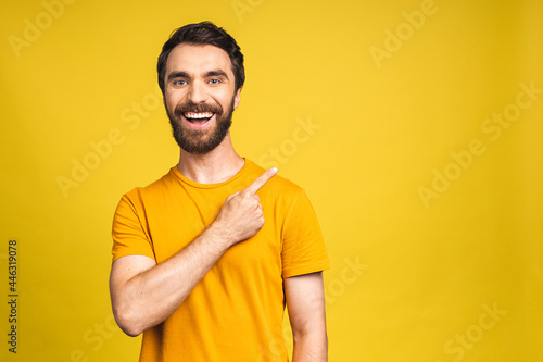 Look over there! Happy young bearded handsome man in casual pointing away up and smiling while standing isolated over yellow background.