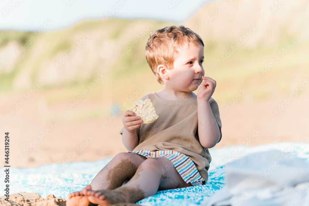 three year old boy eating a snack on the beach in summer at sunset. three year old boy eating a snack on the beach in summer at sunset.