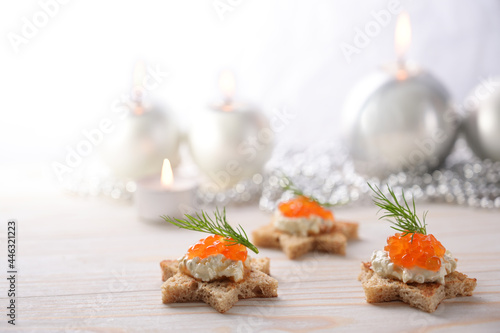Holiday canapes from toasted bread in star shape, cream and red caviar with dill garnish on a light wooden table with candles and festive Christmas decoration, copy space