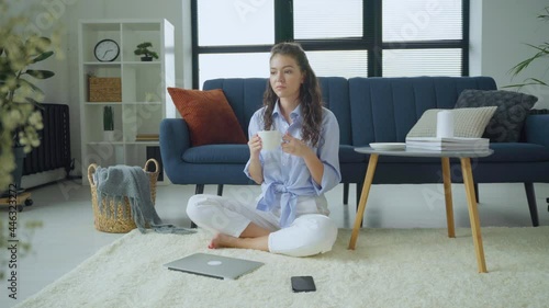 Young caucasian woman sitting on the floor in the living room working on the phone with a laptop. Freelancer is working at home with a laptop.