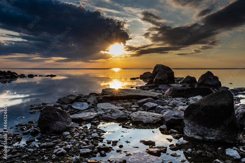 An image of a calm lake with a rocky shore at sunset. The clouds in the sky and the boulders on the beach provide a strong contrast to the warm setting sun at the end of a hot summer day.