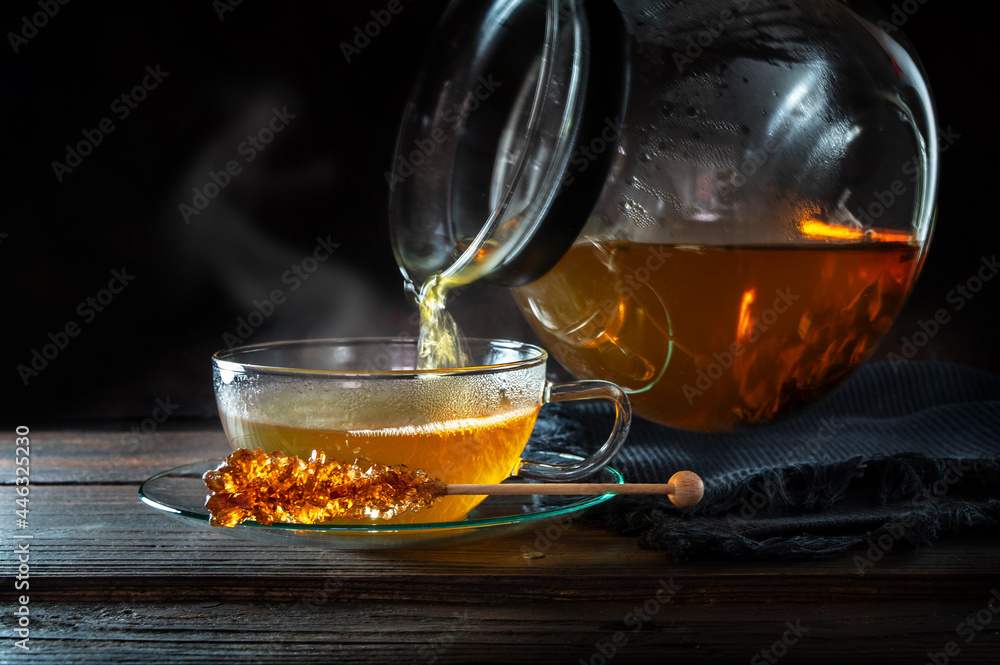 Hot steaming tea is poured from a pot into a glass cup, served with a stick of rock candy, motion blur, dark rustic wooden background with copy space, selected focus Hot steaming tea is poured from a pot into a glass cup, served with a stick of rock candy, motion blur, dark rustic wooden background with copy space, selected focus