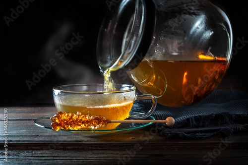 Hot steaming tea is poured from a pot into a glass cup, served with a stick of rock candy, motion blur, dark rustic wooden background with copy space, selected focus