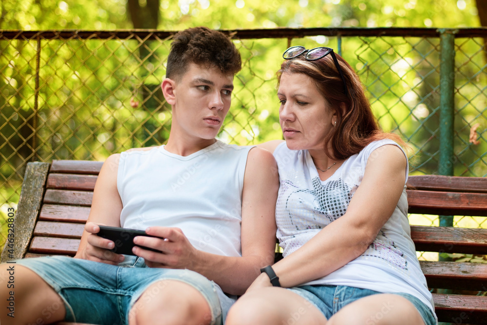 A woman and her teenage son are sitting on a bench in the park and ...