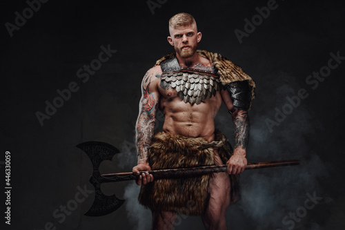 Brutal tattooed warrior wearing light armour and fur holding two-handed axe in dark studio