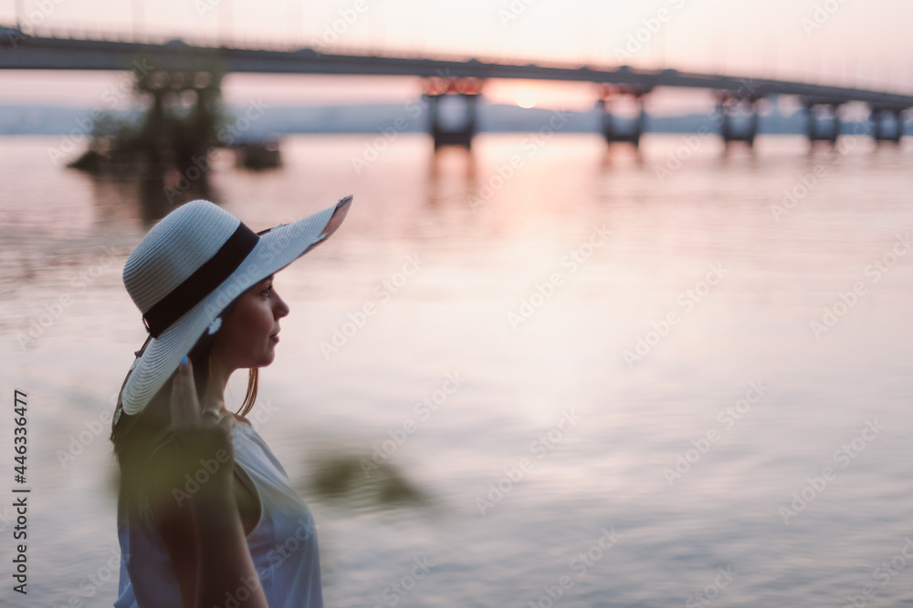 A woman on a river beach at sunset. Profile portrait of a Caucasian woman in a straw hat relaxing and meditating at sunset by the bridge on the river at sunset with the silhouette of the city. 