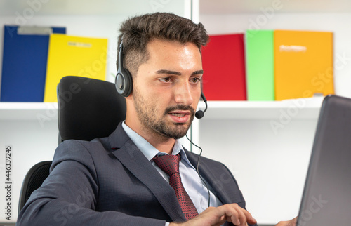 Portrait of businessman, call center agent wearing microphone headset working with laptop in office.  Man in suit consulting client on phone in customer support service. Help, answering and telemarket