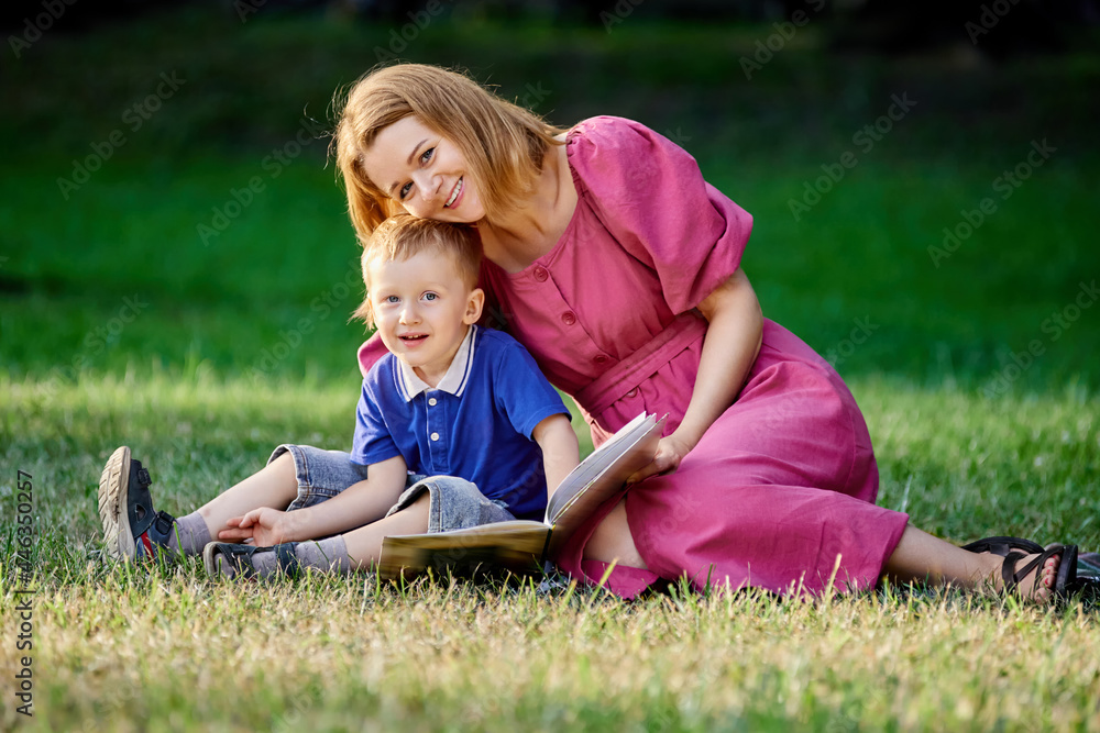 Young smiling woman reads book to little boy sitting on grass in park. Young smiling woman reads book to little boy sitting on grass in park.