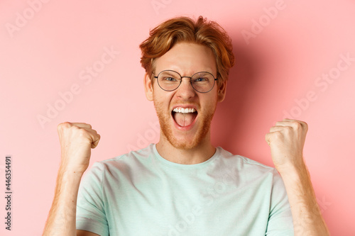 Close up of satisfied lucky redhead man winning, shouting from joy and making fist pump, celebratig victory, standing like champion over pink background photo