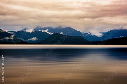 The mountain range bordering Lake Kaniere being reflected on the lake surface on  a dark and rainy wet cloudy moody late afternoon © Stewart