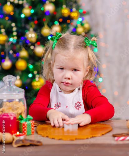 Little cute girl flour sprinkles rolled dough on the background of the Christmas tree