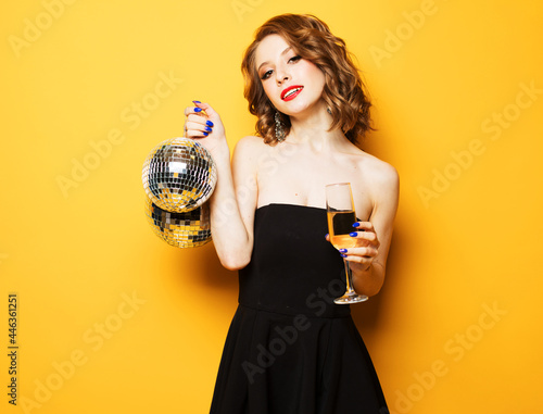 Young blonde woman in evening black dress holding a glass of champagne and disco ball. Brighrt make up and Wavy hairstyle. Over yellow background. photo