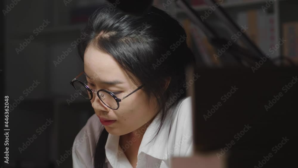 Close up tired young Asian woman wearing glasses, working hard till late night, checking stock and taking a note in working room at home, counting order, taking glasses off and touching head Close up tired young Asian woman wearing glasses, working hard till late night, checking stock and taking a note in working room at home, counting order, taking glasses off and touching head