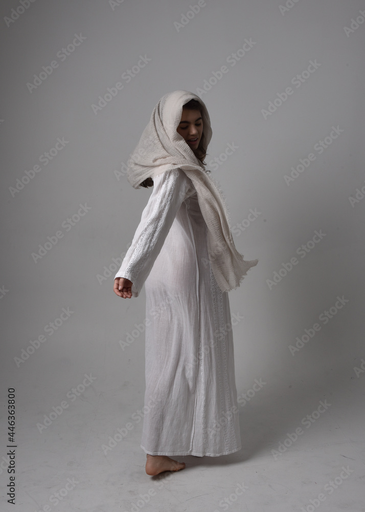 Full length portrait of young woman wearing classical white gown and a head covering veil in biblical style, standing pose on light studio background.