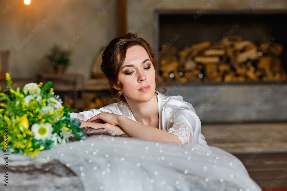 Naklejka premium A beautiful young brunette in a silk shirt sits next to the bed with a bouquet of flowers and a wedding veil. Bride's morning
