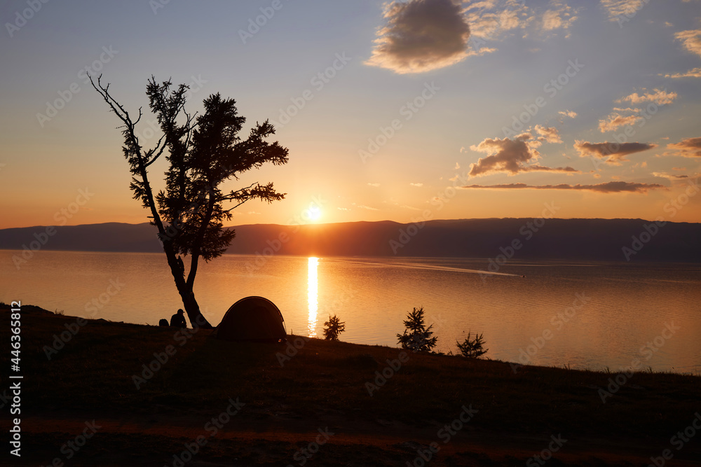 Beautiful sunset on the lake. A lonely man is seated on the shore and admiring the sunset. There is a tent nearby. 