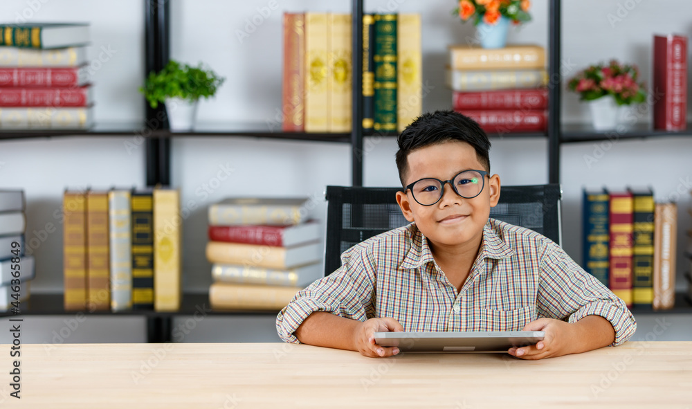 Fototapeta premium Smart Asian boy wearing glasses and grey checked shirt happily sitting in reading room and enjoy study of searching funny info by tablet on desk