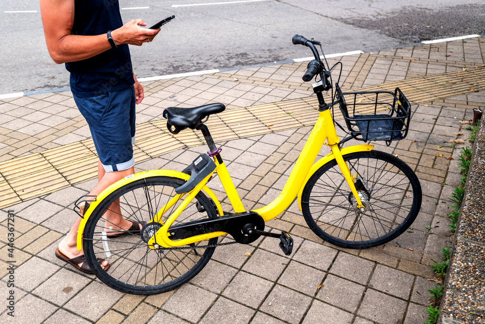 Naklejka premium BIKE YELLOW and mobile phone. man using e vehicle rent service with smartphone in urban city street and park. Young man rent an eco transportation in summer