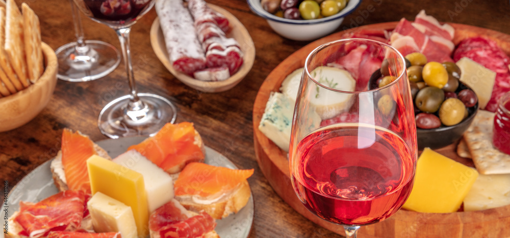 Gourmet wine appetizers panorama with a glass of rose wine, a cheese and charcuterie board, and salmon sandwiches. Italian antipasti or Spanish tapas panoramic banner for a menu