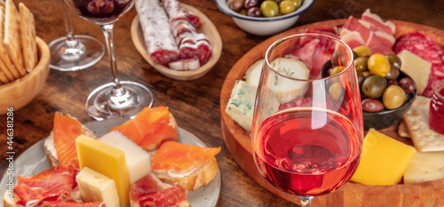 Gourmet wine appetizers panorama with a glass of rose wine, a cheese and charcuterie board, and salmon sandwiches. Italian antipasti or Spanish tapas panoramic banner for a menu