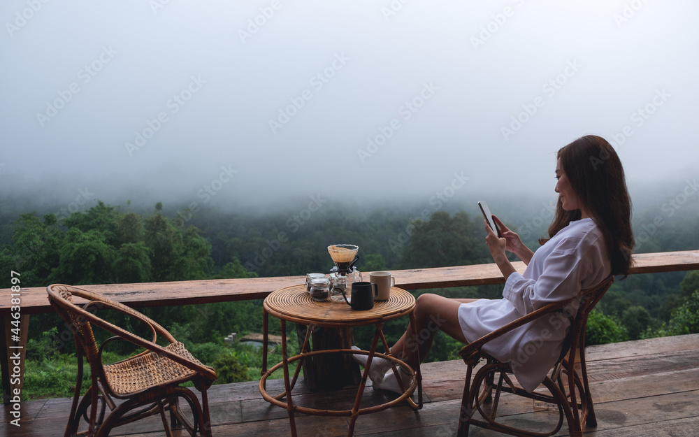 A young asian woman using mobile phone while sitting on balcony with a beautiful nature view on foggy day