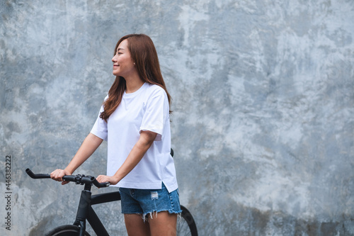 Portrait image of a beautiful young asian woman wearing white t-shirt and jean short pant with bicycle and concrete wall background
