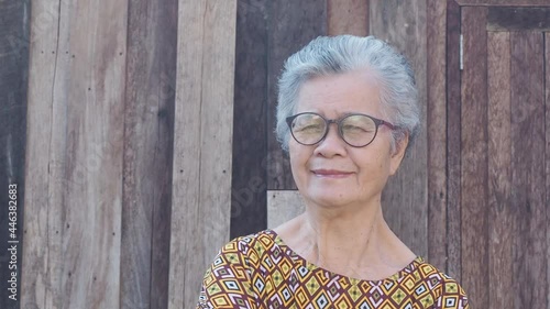 Portrait of an eldelry Asian woman with short gray hair, smiling and looking at the camera while standing outdoors with wooden wall background. Concept of aged people and healthcare.
