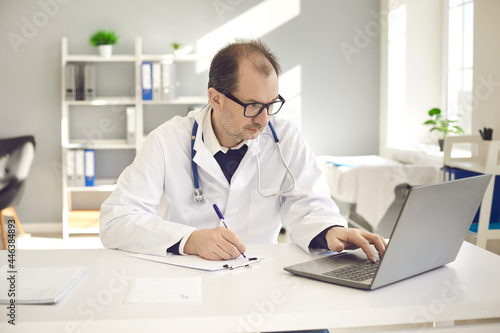 Serious man doctor in white uniform with stethoscope using laptop writing notes in medical journal patient card sitting at desk in clinic office. Professional remote consultation, online medicine