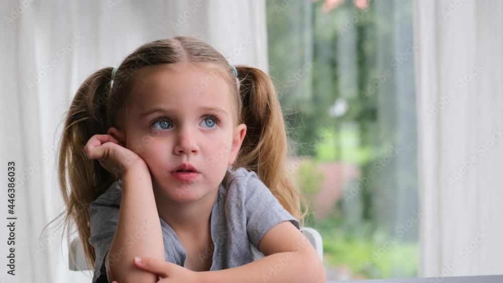 Bored preschooler girl blonde with long pigtails puts head on hands and looks straight sitting at home against window close view. Child holds head with hand and looks up and away dreaming