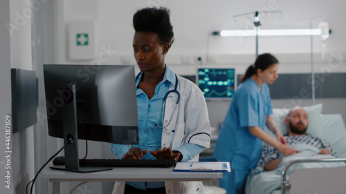 Front view of afro american doctor typing medical expertise on computer while in background nurse checking heartbeat pulse using oximeter. Hospitalized patient having respiratory disorder