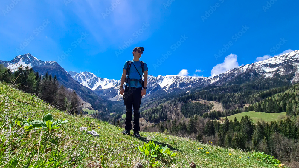 A man with a big hiking backpack enjoying the panoramic view on Baeren Valley in Austrian Alps. The highest peaks are snow-capped. Lush green pasture. Clear and sunny day. High mountain chains.