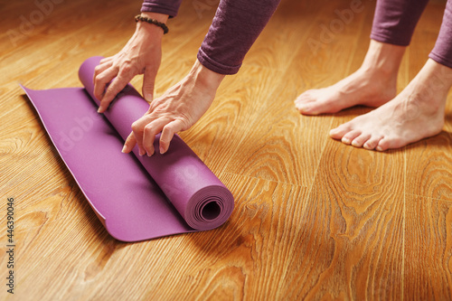 A woman's hands fold a lilac yoga or fitness mat after a workout at home in the living room.