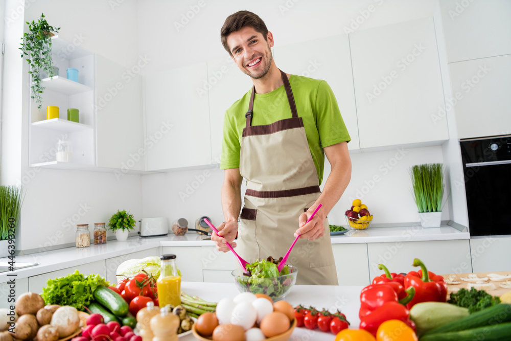 Portrait of attractive cheerful guy cooking delicious fresh salad dish culinary hobby free time at home kitchen indoors