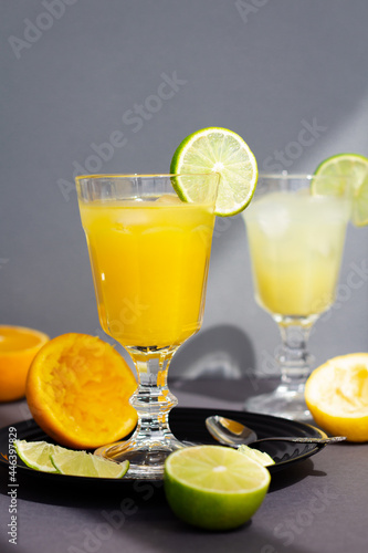 a glass of refreshing citrus drink or lemonade or alcoholic cocktail, a pair of vintage goblets among sliced lemons, oranges and limes