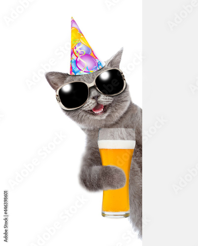 Happy cat wearing birthday cap holds glass of beer and look from behind empty board. isolated on white background