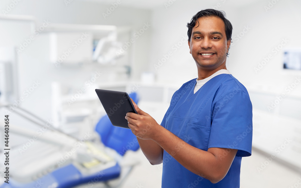 medicine, stomatology and technology concept - happy smiling indian doctor or male dentist in blue uniform using tablet pc computer over dental clinic office background