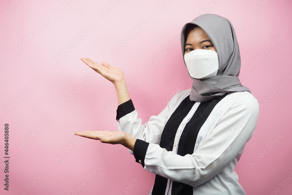 Muslim woman wearing white mask, with hand pointing to empty space presenting something, isolated on pink background Muslim woman wearing white mask, with hand pointing to empty space presenting something, isolated on pink background