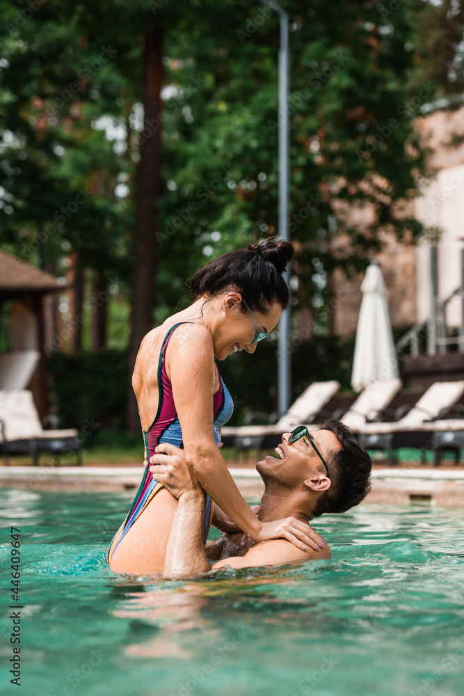 Side view of positive man rising cheerful girlfriend in swimming pool during vacation