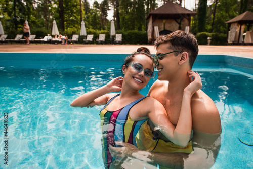 Smiling man in sunglasses embracing girlfriend with closed eyes in swimming pool