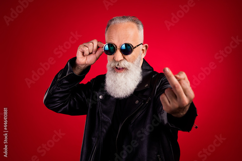Portrait of attractive fashionable elderly grey-haired man showing middle finger touching specs isolated over bright red color background photo